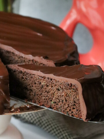 A slice of keto zucchini chocolate cake with low carb ganache on a server being pulled out of the cake on a white pedestal.