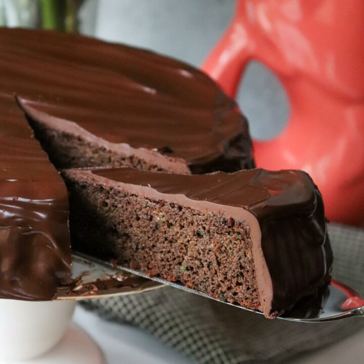 A slice of keto zucchini chocolate cake with low carb ganache on a server being pulled out of the cake on a white pedestal.
