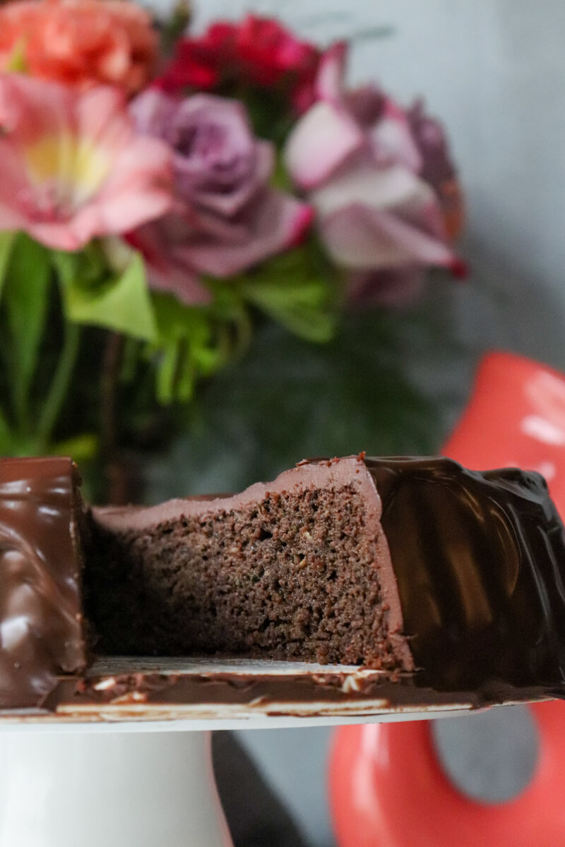 A piece taken out of a keto chocolate zucchini cake on a decorative white pedestal.