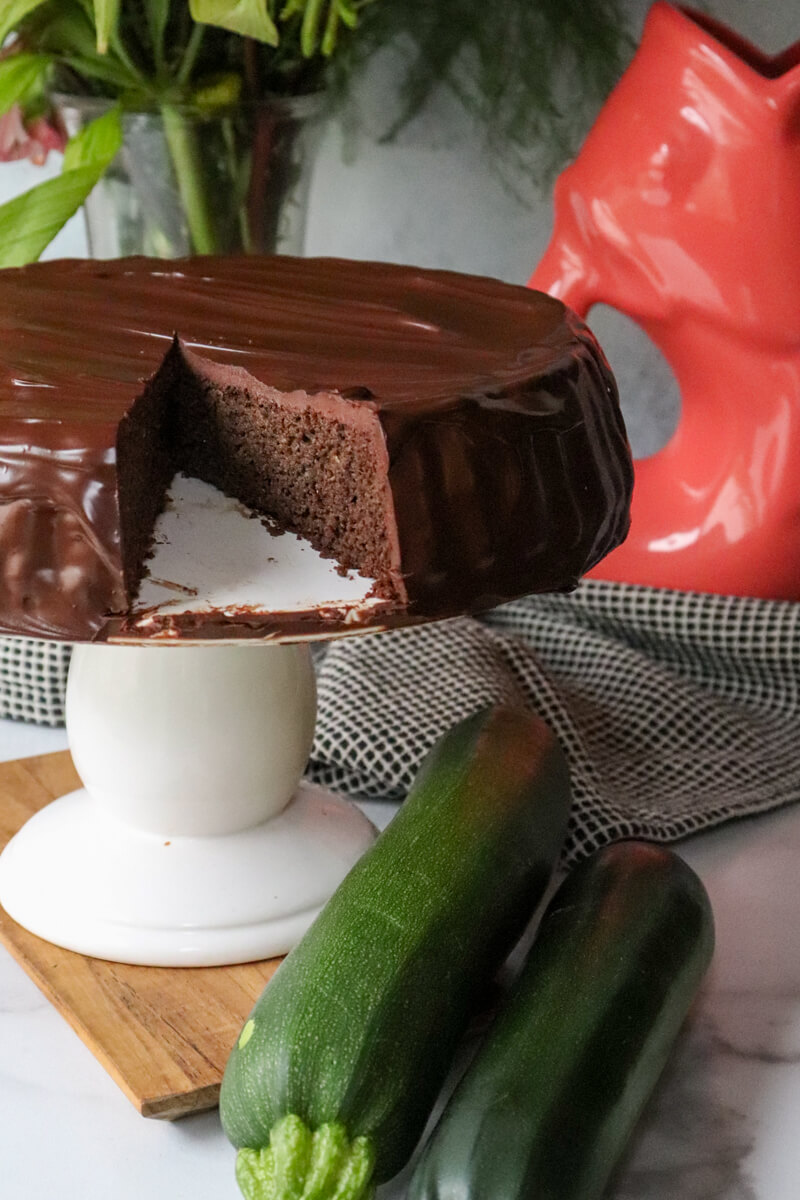 A piece taken out of a keto chocolate zucchini cake on a decorative white pedestal.
