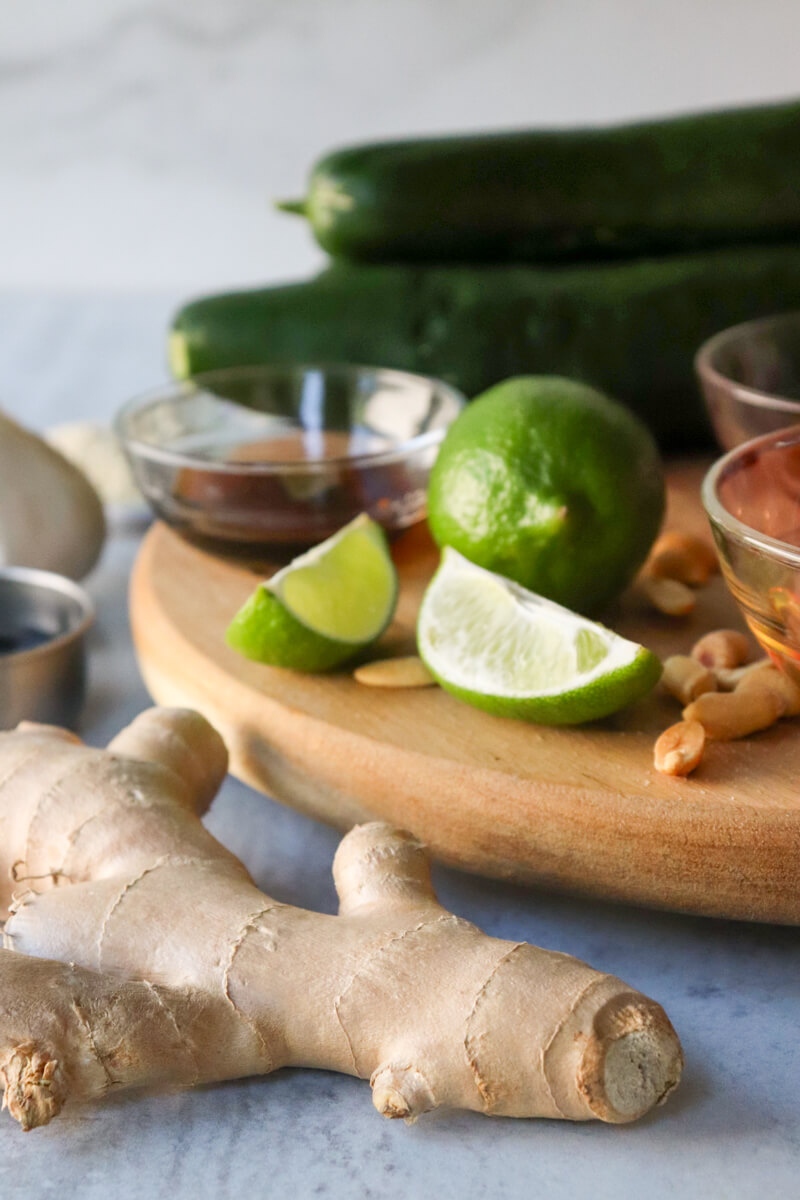 Front view of a ginger root next to a circle board with lime, peanuts, cucumbers, and ingredients for the dressing.