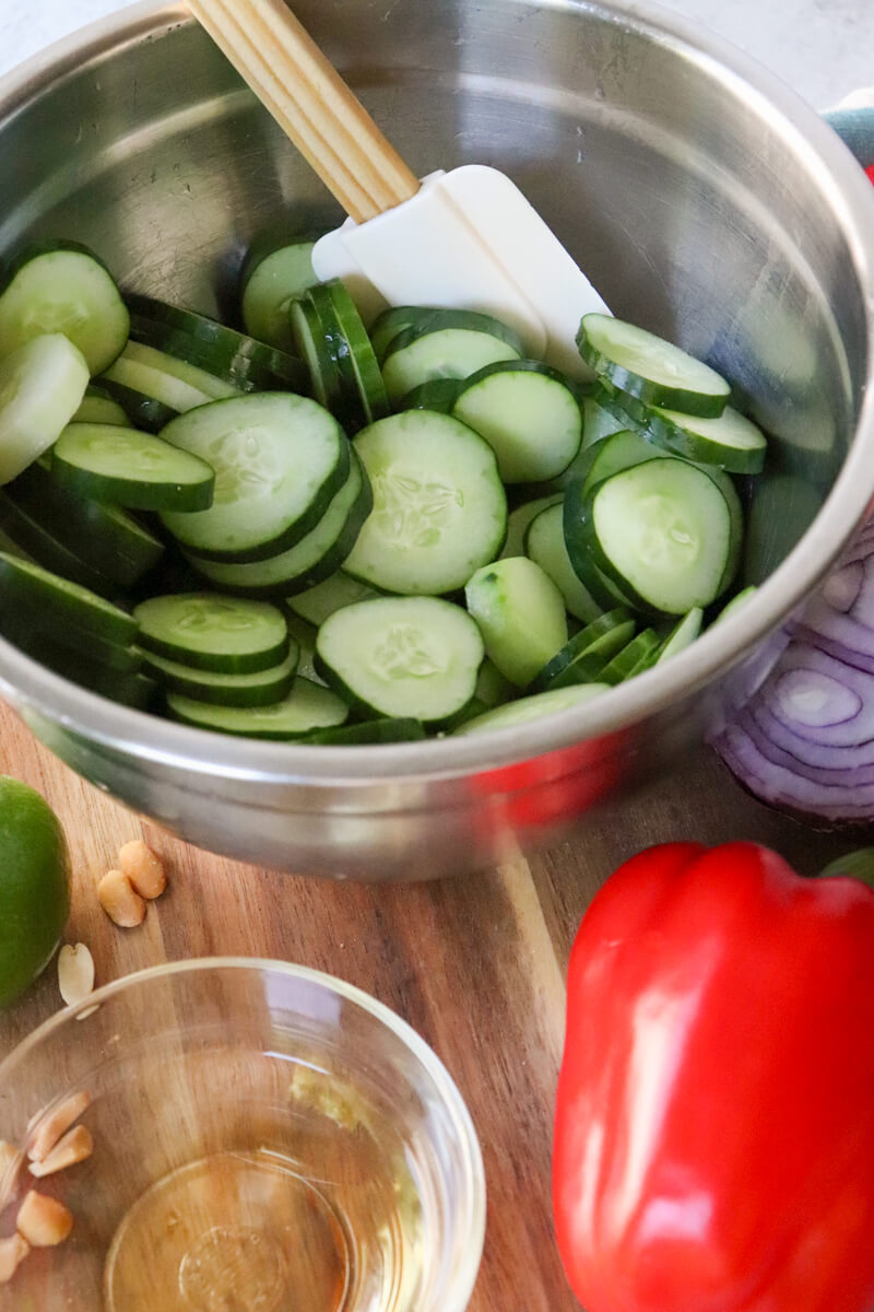 A silver bowl of sliced cucumbers next to a red pepper and red onion.