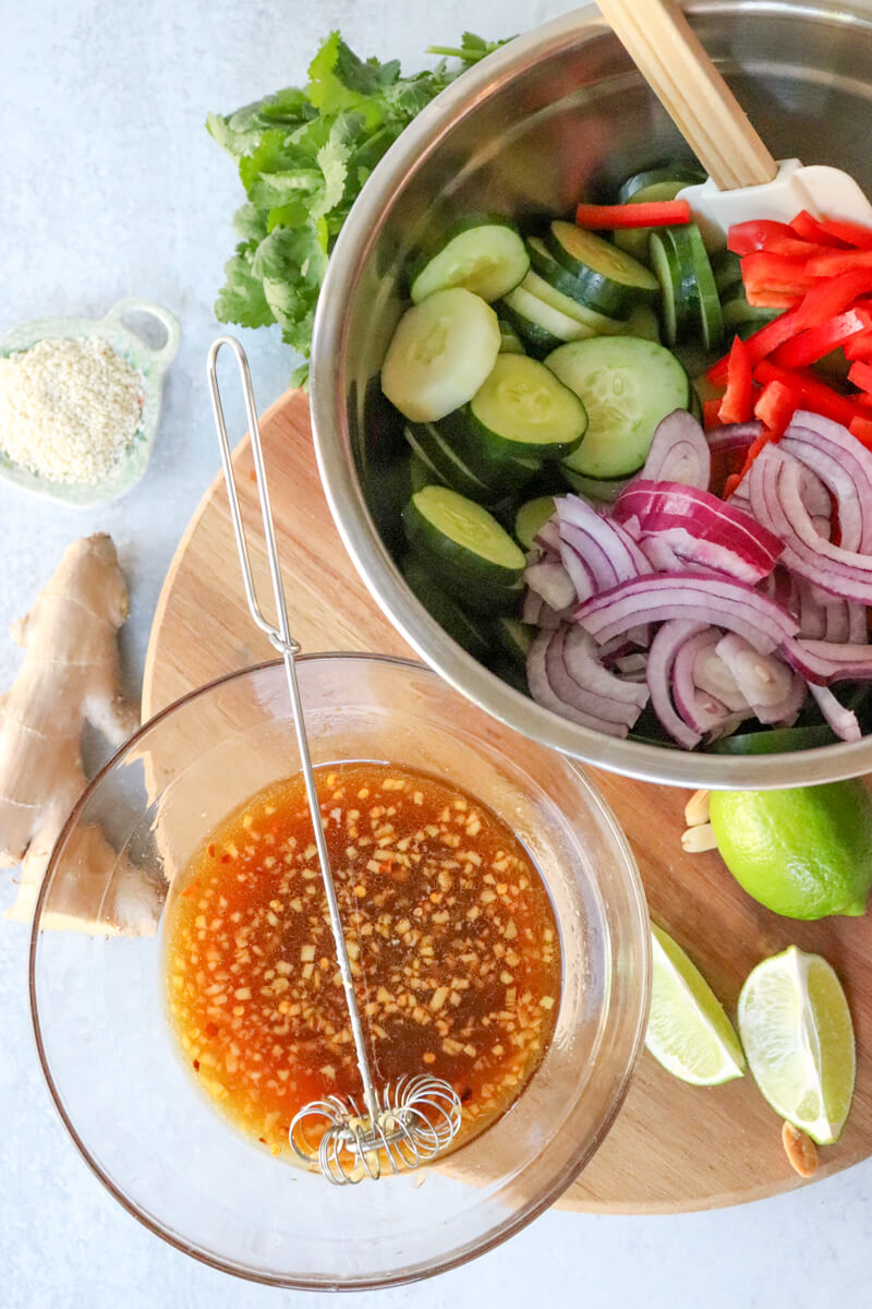 Overhead view of a silver bow with the cucumber mixture and a glass bowl with the dressing. All on a board and lime wedges.