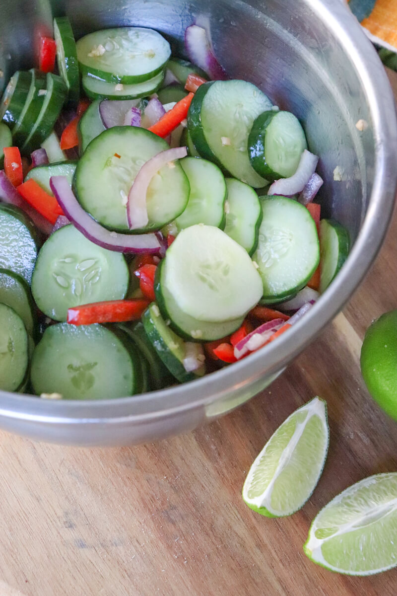 A silver bowl of mixed cucumbers, red onion, and red pepper on a board with lime wedges.
