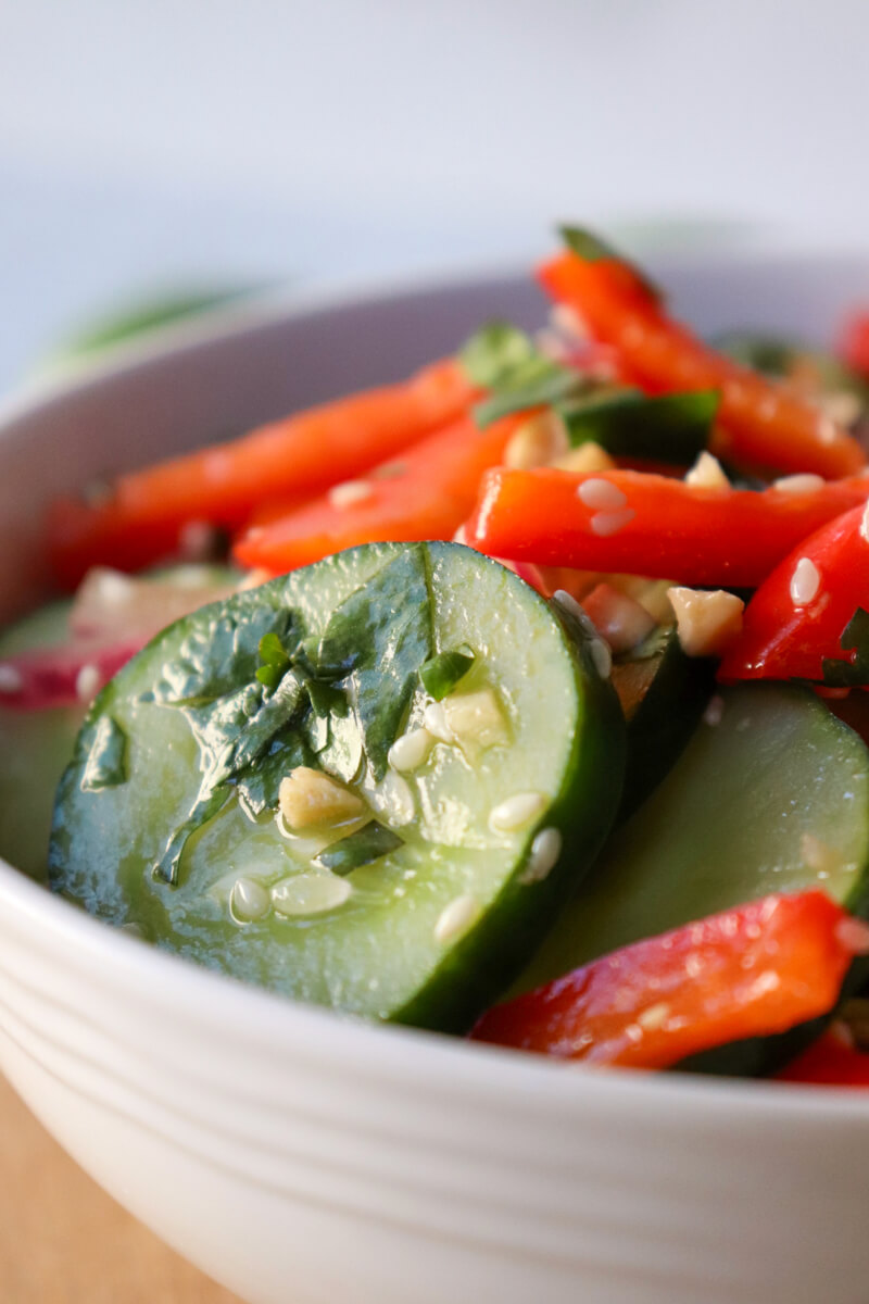 A close-up view of a white bowl of Thai cucumber salad with cucumbers, red peppers, and red onion, dotted with sesame seeds and cilantro.