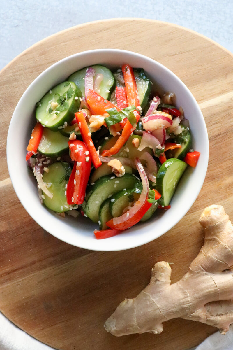 Overhead view of a white bowl of Thai cucumber salad with cucumbers, red peppers, and red onion, dotted with sesame seeds and cilantro. Sitting on a wooden circle board with ginger root.