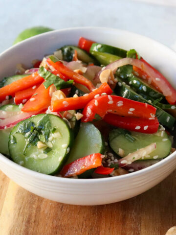 A white bowl of Thai cucumber salad with cucumbers, red peppers, and red onion, dotted with sesame seeds and cilantro. Sitting on a wooden circle board.