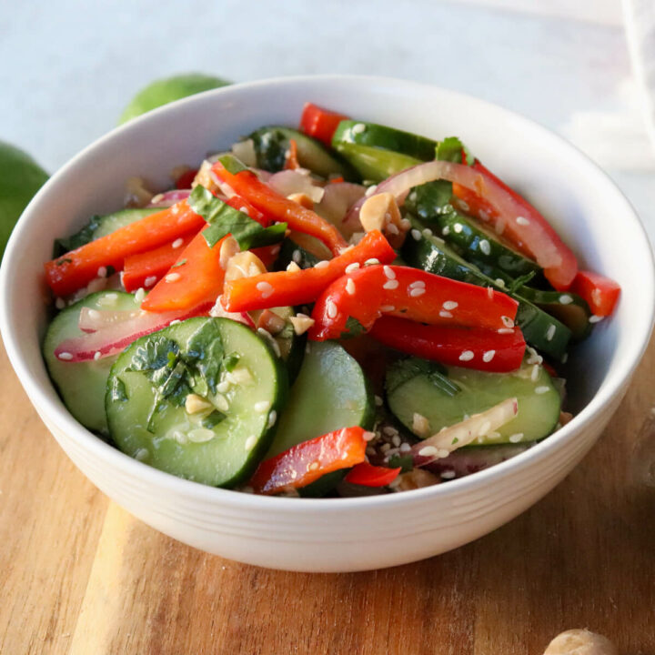 A white bowl of Thai cucumber salad with cucumbers, red peppers, and red onion, dotted with sesame seeds and cilantro. Sitting on a wooden circle board.