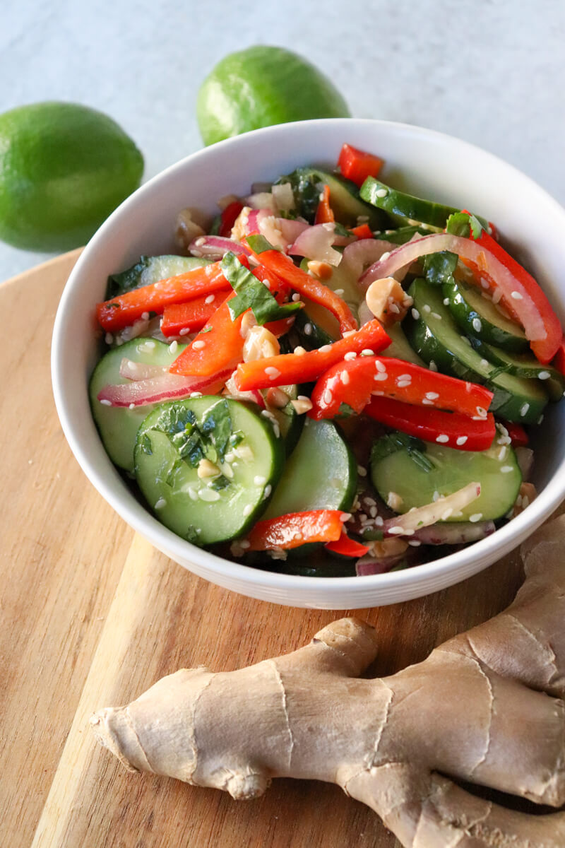 A white bowl of Thai cucumber salad with cucumbers, red peppers, and red onion, dotted with sesame seeds and cilantro. Sitting on a wooden circle board with a ginger root in front.