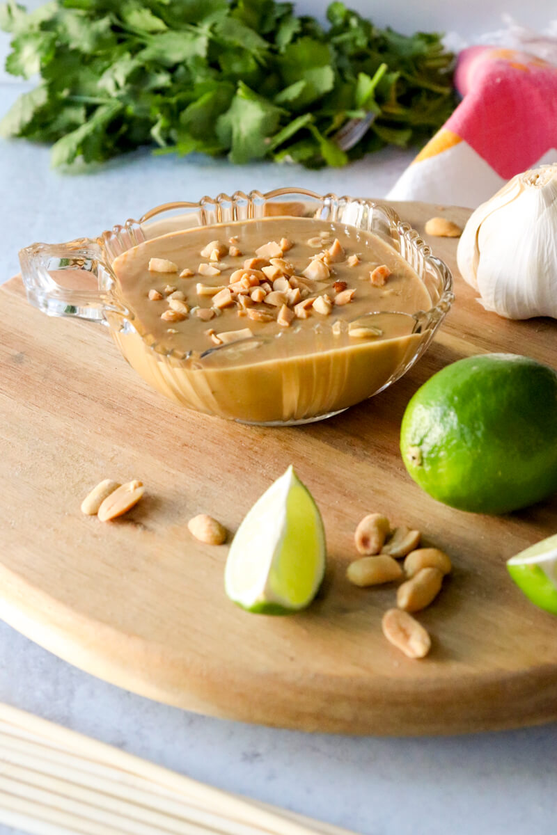 A glass bowl of low carb peanut sauce on a wooden lazy Susan. Cilantro is in the background with lime wedges and peanuts in front of the sauce.