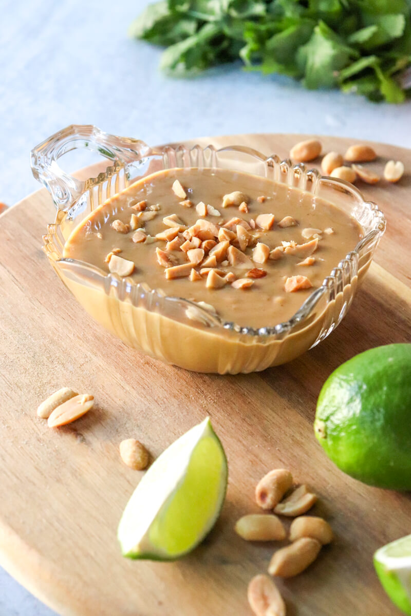 A glass bowl of low carb peanut sauce on a wooden lazy Susan. Cilantro is in the background with lime wedges and peanuts in front of the sauce.
