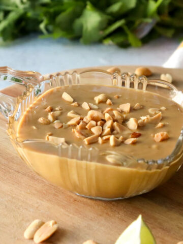 A glass bowl of low carb peanut sauce on a wooden lazy Susan. Cilantro is in the background with lime wedges and peanuts in front of the sauce.