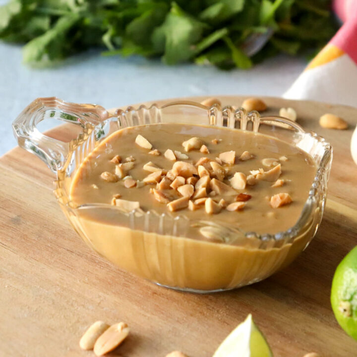 A glass bowl of low carb peanut sauce on a wooden lazy Susan. Cilantro is in the background with lime wedges and peanuts in front of the sauce.