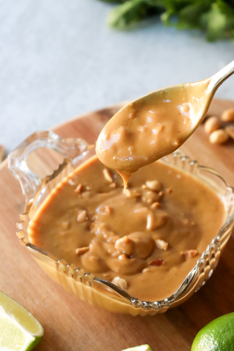 A close up of a glass bowl of low carb Thai peanut sauce sprinkled with extra peanuts, and a spoon lifting some out to drizzle over something