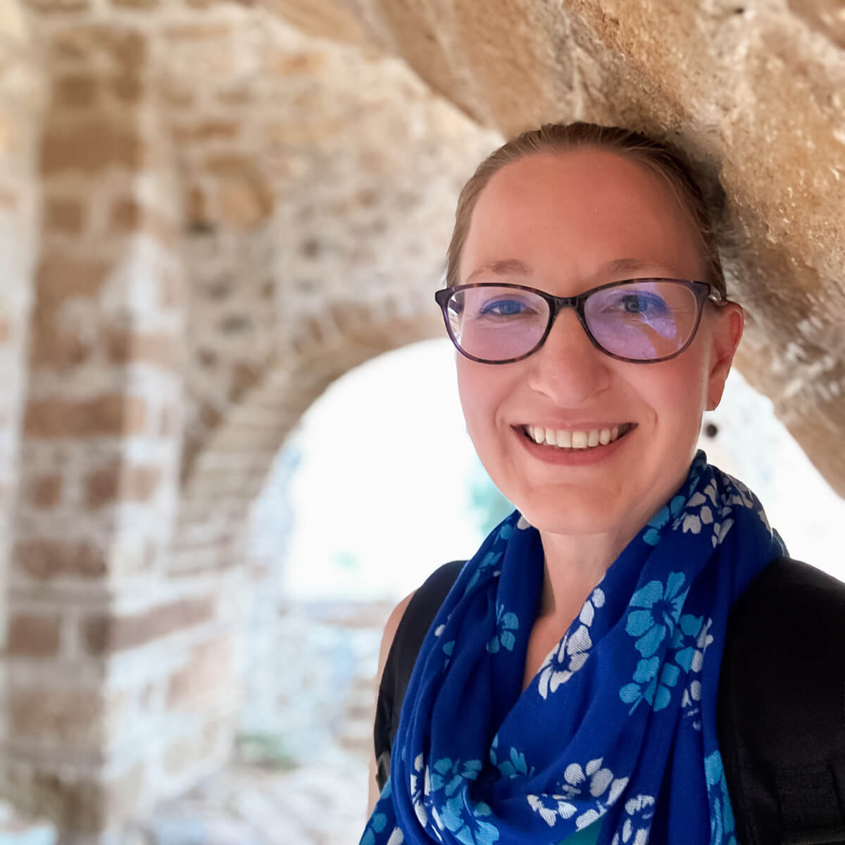 Headshot of Lauren wearing a blue scarf with hair pulled back under a stone archway in Monemvasia, Greece.
