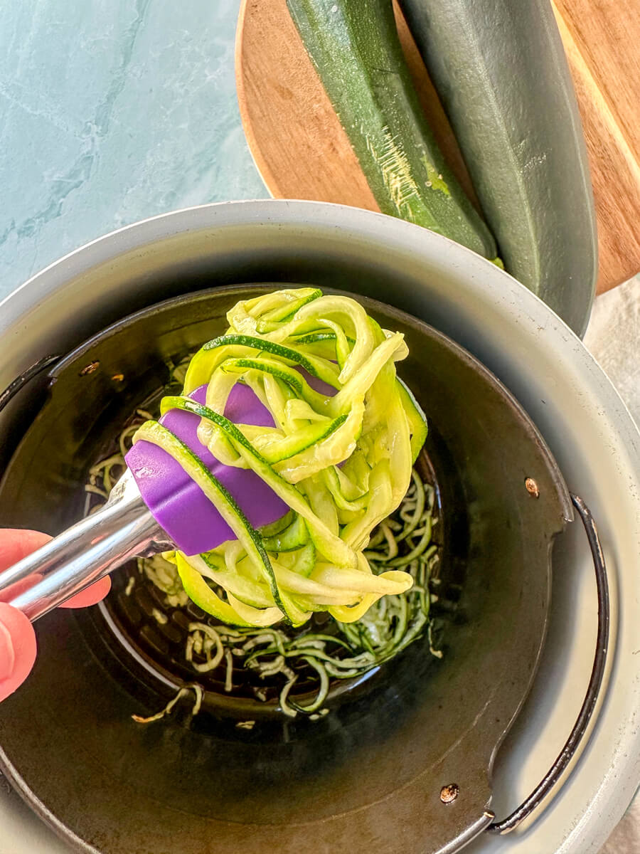 Overhead view of air fried zoodles in the metal air fryer basket with tongs holding some up to view
