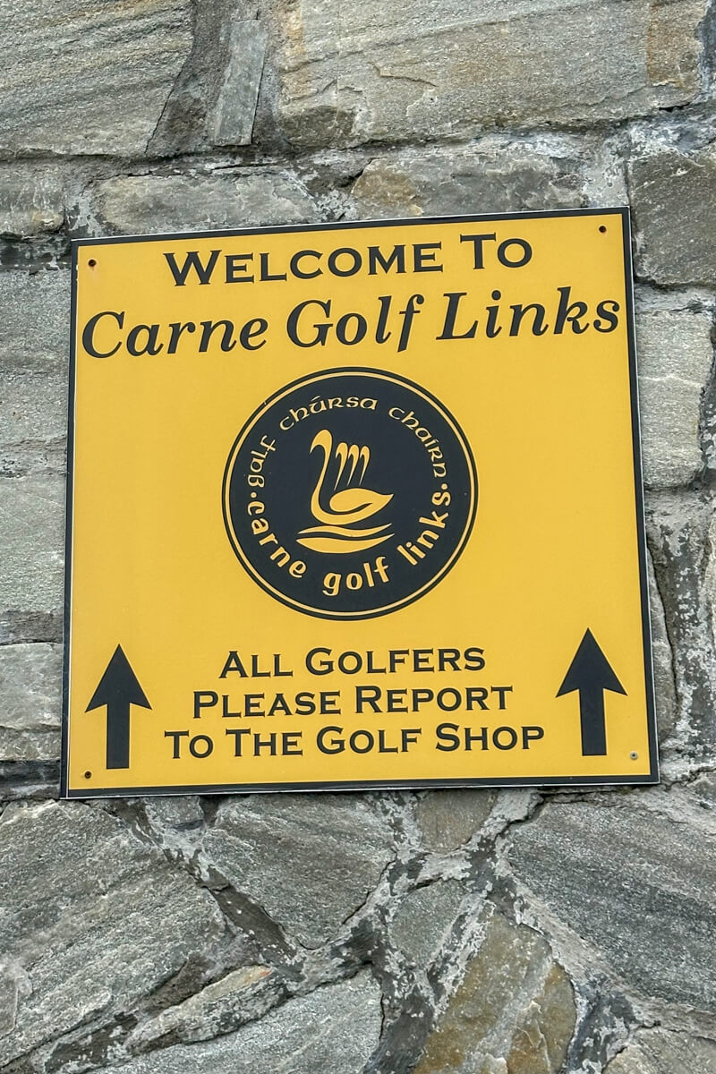 A tan/yellow Welcome to Carne Golf Links sign on a stone wall