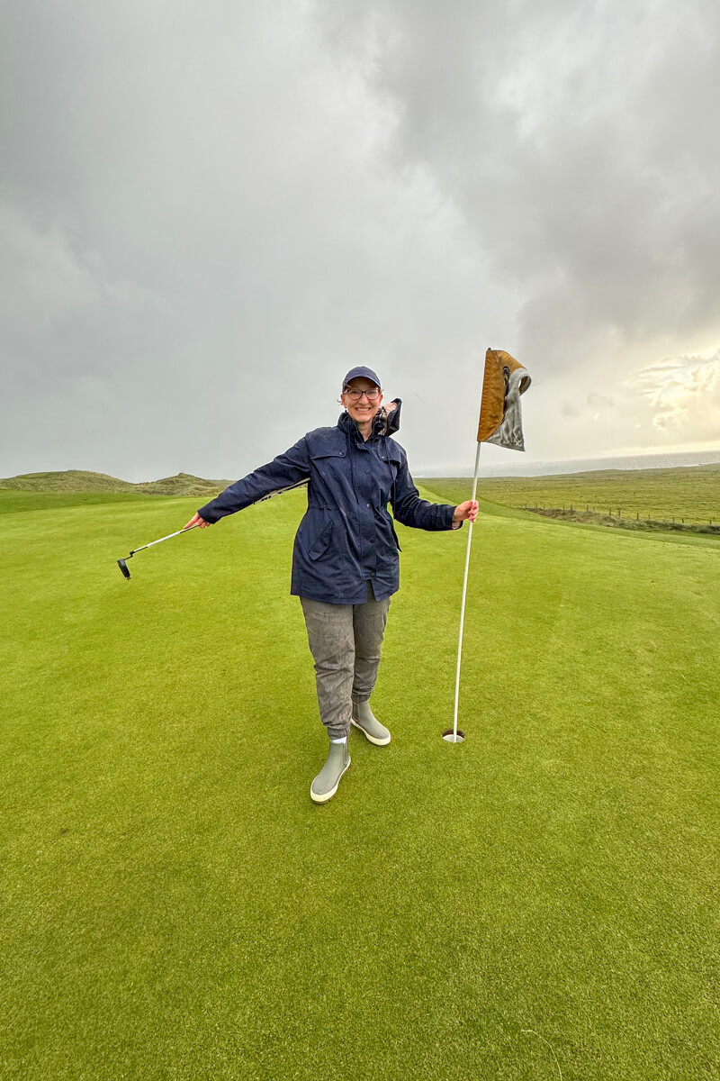 Lauren holding out a golf club standing with the flag in hand by the hole at Carne Golf Links #3