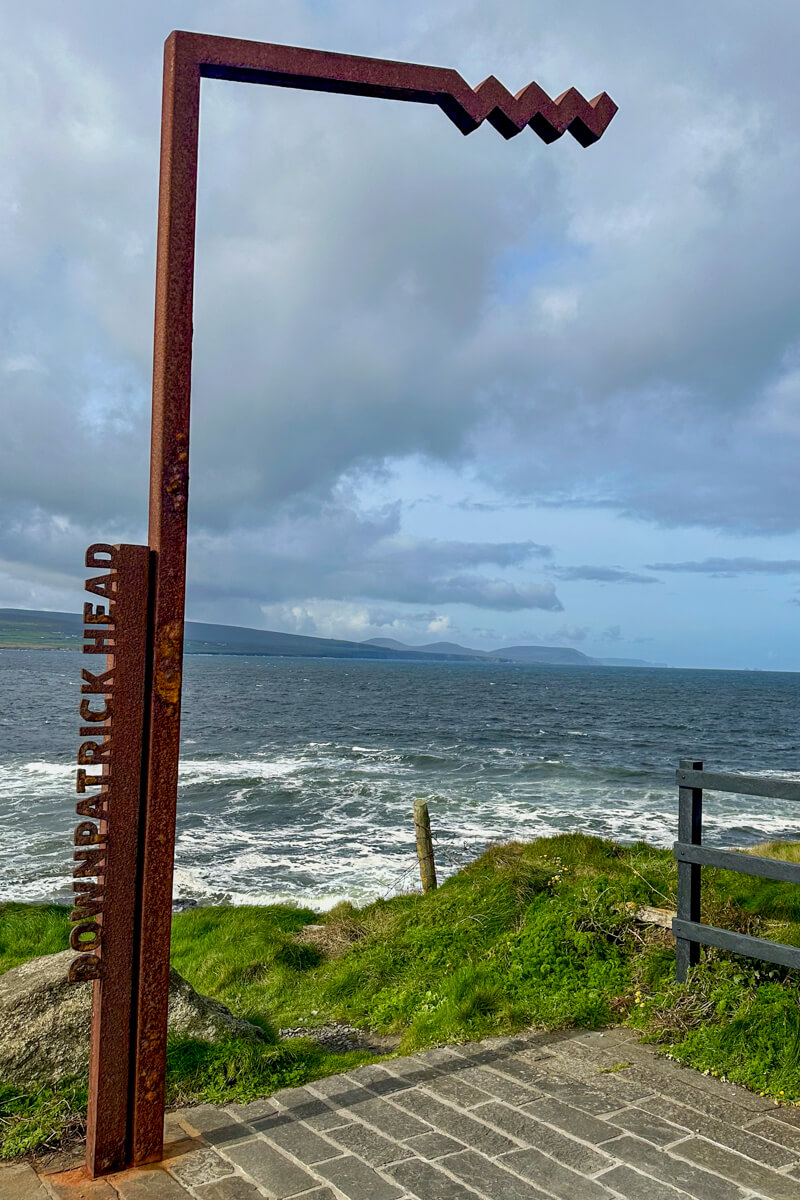 Metal entrance sign with the beach and water in the distance to Downpatrick Head.