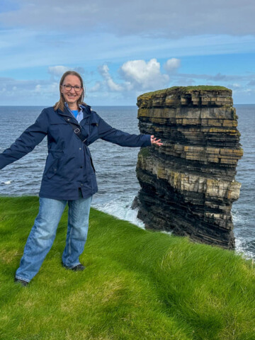 Lauren in jeans and a navy rain jacket standing on the edge of a green cliff in front of the Dún Briste sea stack at Downpatrick Head.