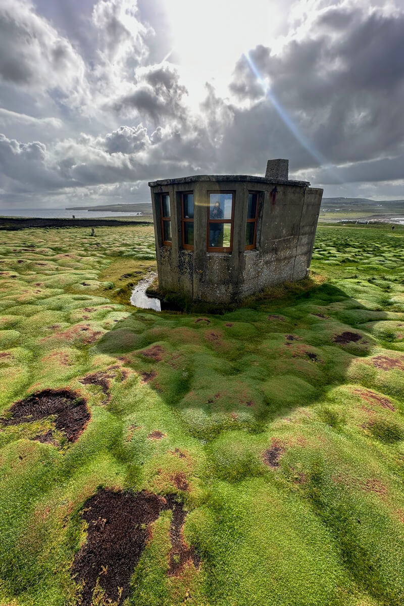 A small gray building on the distinctive bright green grass of Downpatrick Head with sunlight coming down.