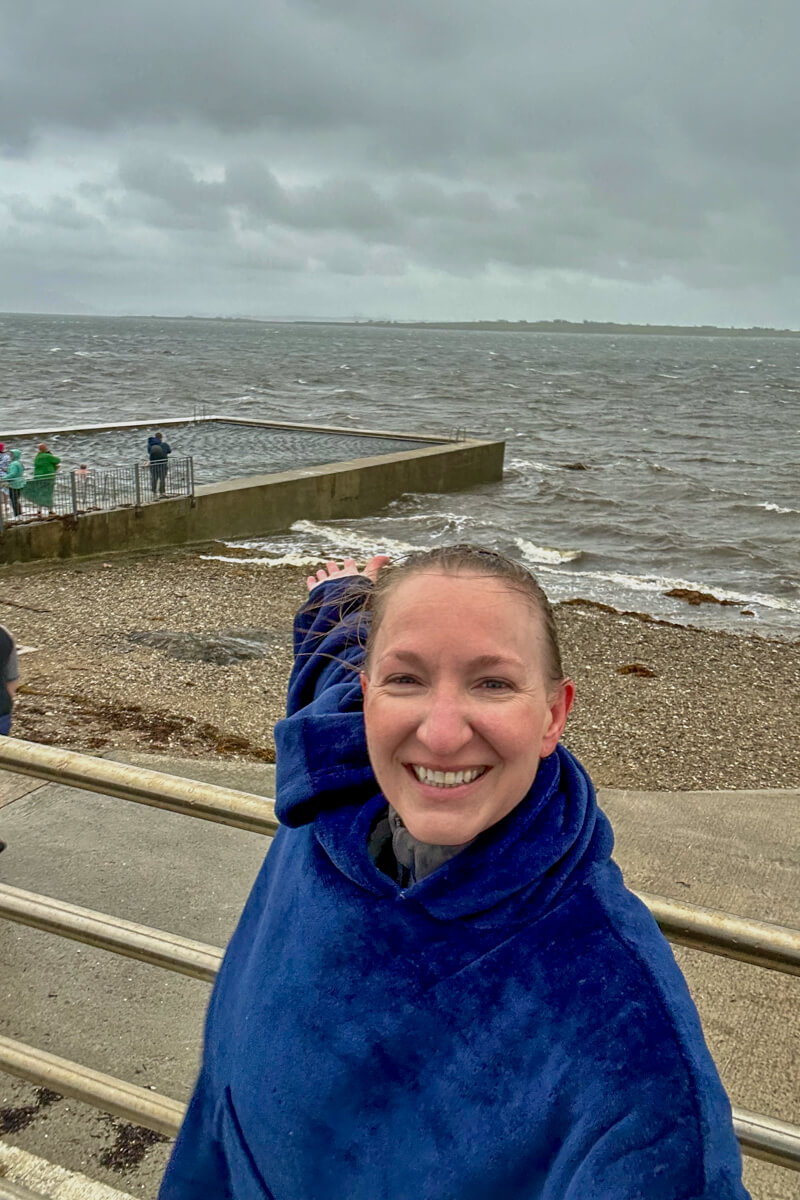 Lauren smiling wearing a blue swimming rode pointing back at the Belmullet Tidal Pool.