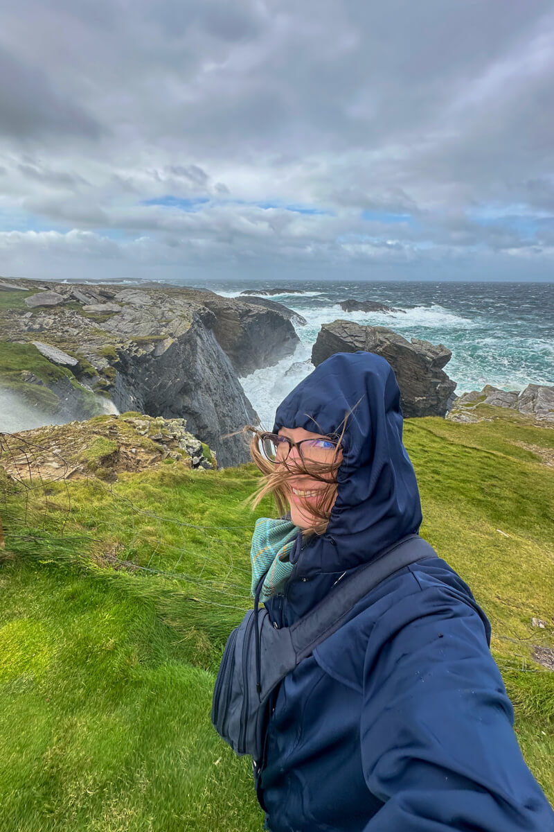 Lauren in a blue raincoat with a green scarf and hood up with hair blowing pointing to a gorgeous view of cliffs, rocks, and waves.