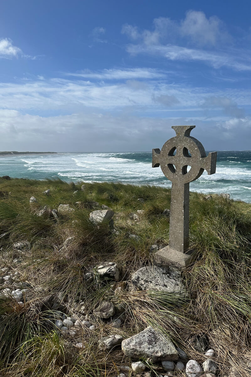 A Celtic cross and grave on w grassy hill in front of the beach and Atlantic Ocean at Cross Cemetery in Mayo.