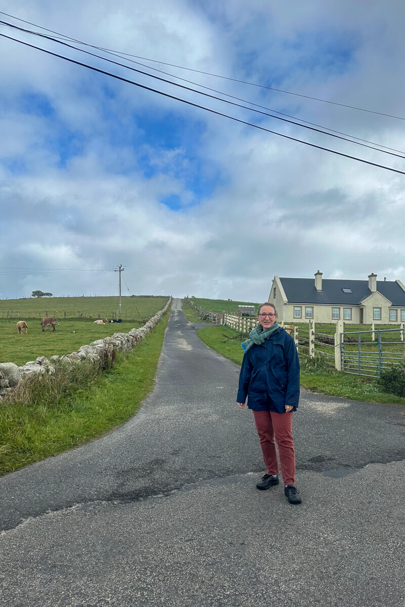 Lauren standing on the street in Fallmore, Ireland with a stone fence on the left and a house with two chimneys on the back right.