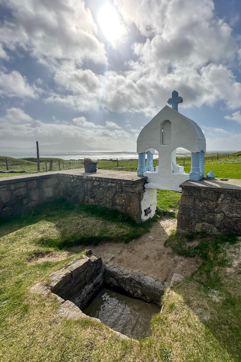 St. Deirbhile's Spring, a square area with water flowing. Surrounded by a stone fence and a white and blue cross marker.