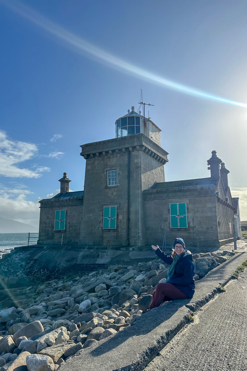 Lauren sitting in front of the Blacksod Lighthouse in north Mayo with her hand reached out toward it.