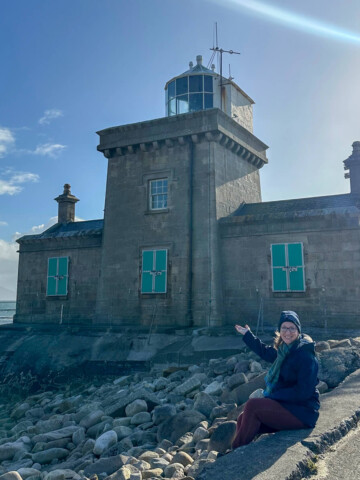 Lauren sitting in front of the Blacksod Lighthouse in north Mayo with her hand reached out toward it.