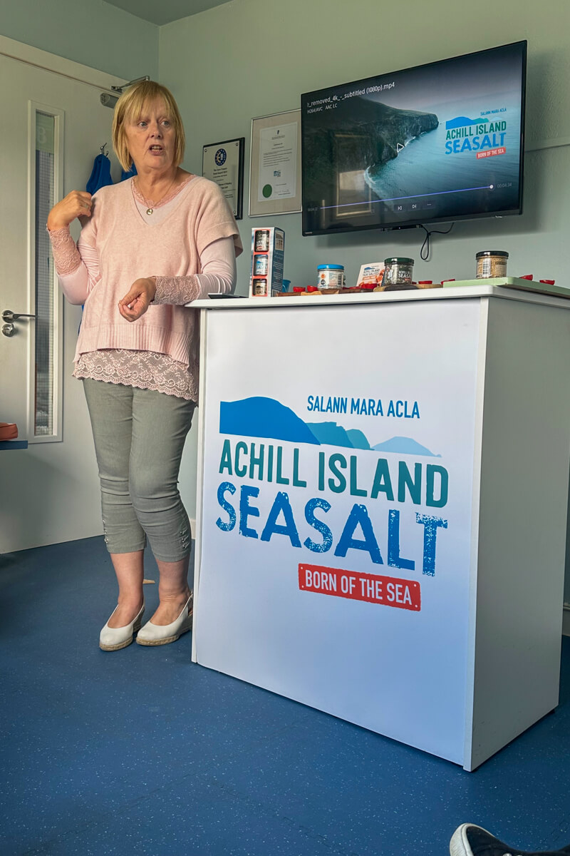 A woman in a pink blouse speaking next to a podium that says Achill Island Seasalt