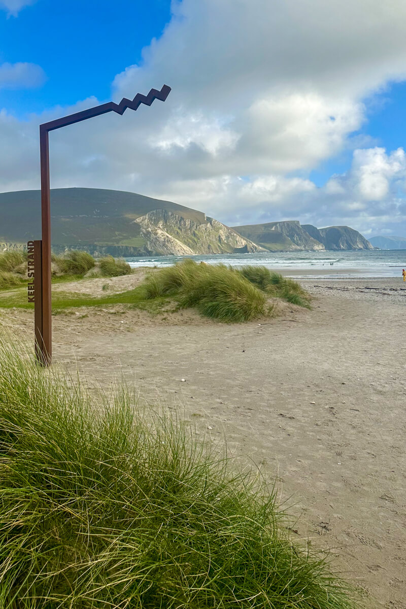 The metal sign and view of Keel Beach sand, water and hills in the background with sun and white clouds