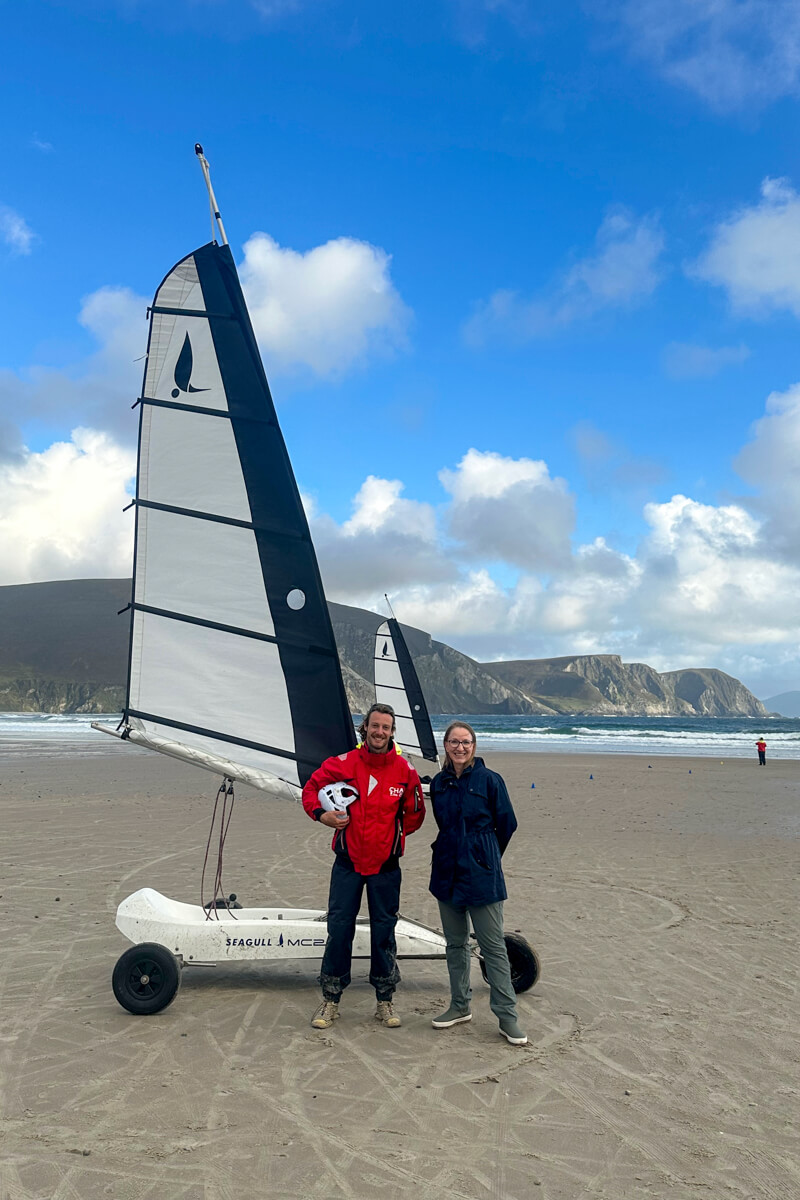 A man in a red jacket standing next to Lauren in her navy raincoat in front of the land sail on Keel Beach