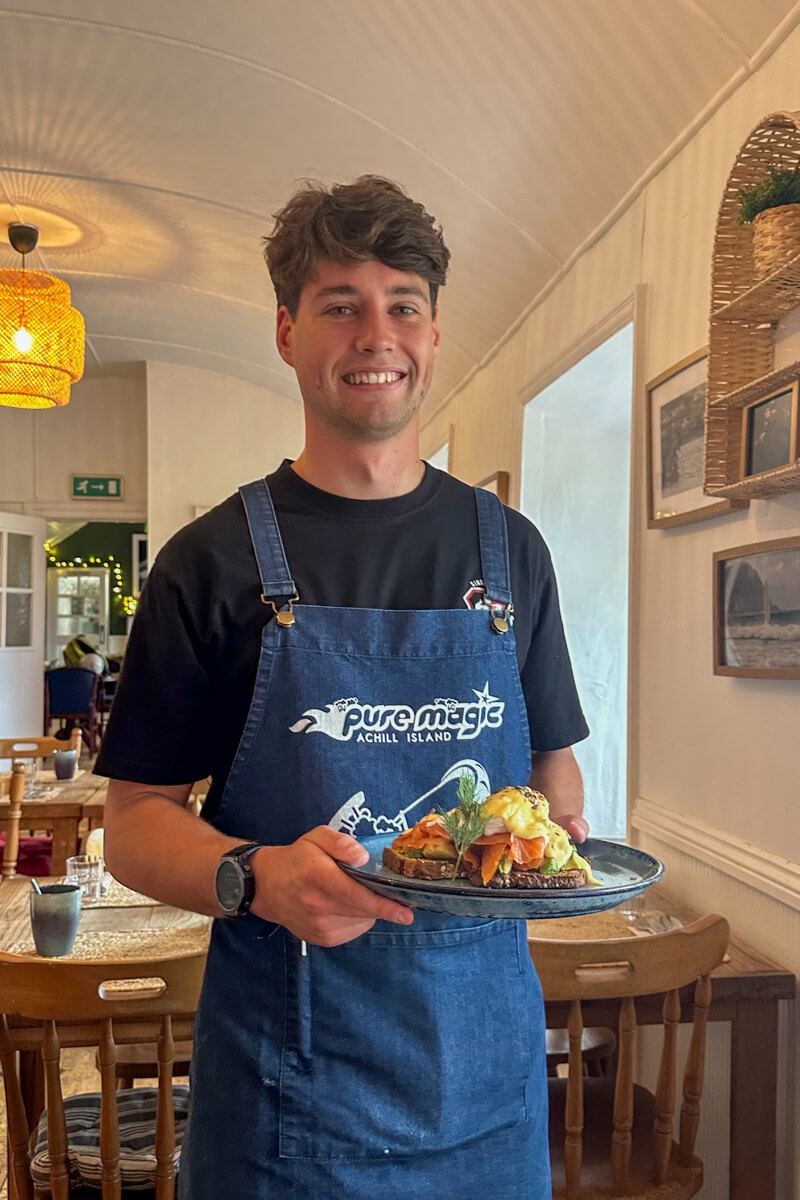 The cheerful waiter in a blue apron bringing breakfast at Pure Magic.