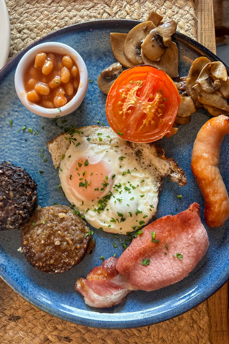 Overhead view of my mini Irish breakfast on a blue plate. Beans, mushrooms, grilled tomato, fried egg, sausage, bacon, blood sausage.