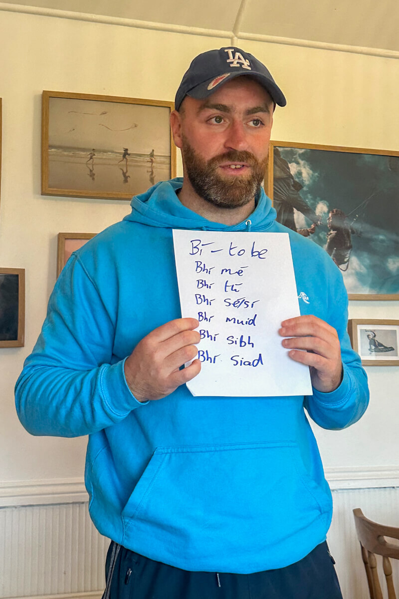Toilach in a light blue sweatshirt and LA Dodgers baseball cap holding up a sign with the conjugation of to be (past tense) in Irish language.