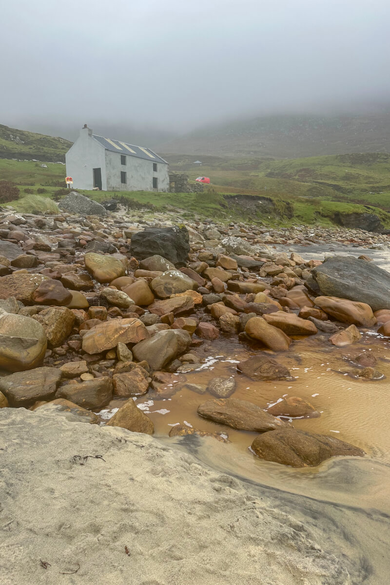 Keem Beach red rocks with a white cottage in the mist and a green and brown hillside.