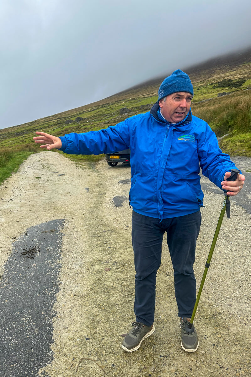 A man in a blue coat and hat and walking stick pointing to the trail to the deserted village on Achill Island
