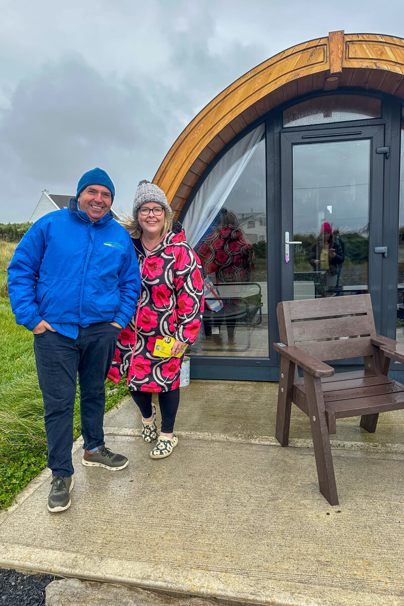 A man in a blue jacket and hat next to a woman in a pink floral knee length coat standing in front of one of Nanny Goose's glamping pods
