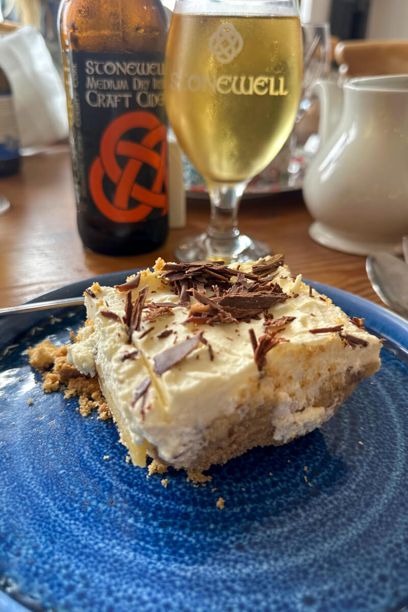 A front view of a piece of banoffee pie on a blue plate with a glass and bottle of cider in the background