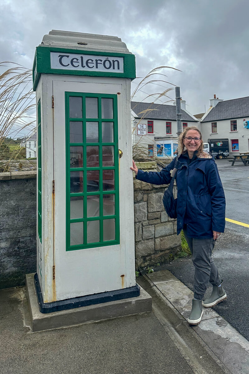 Lauren in a raincoat an green boots standing at the white and green Irish phone booth