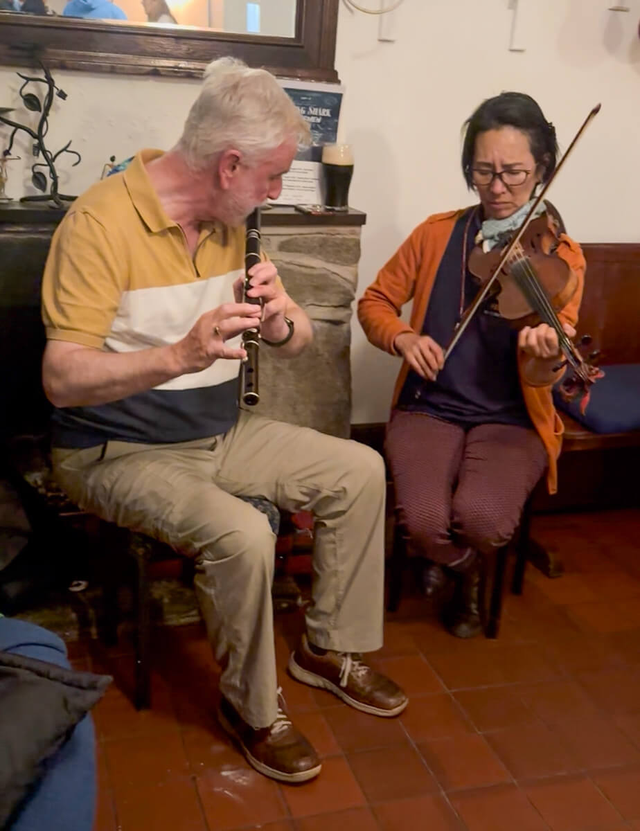 A man playing a wooden flute and a woman in classes and an orange sweater playing a violin.