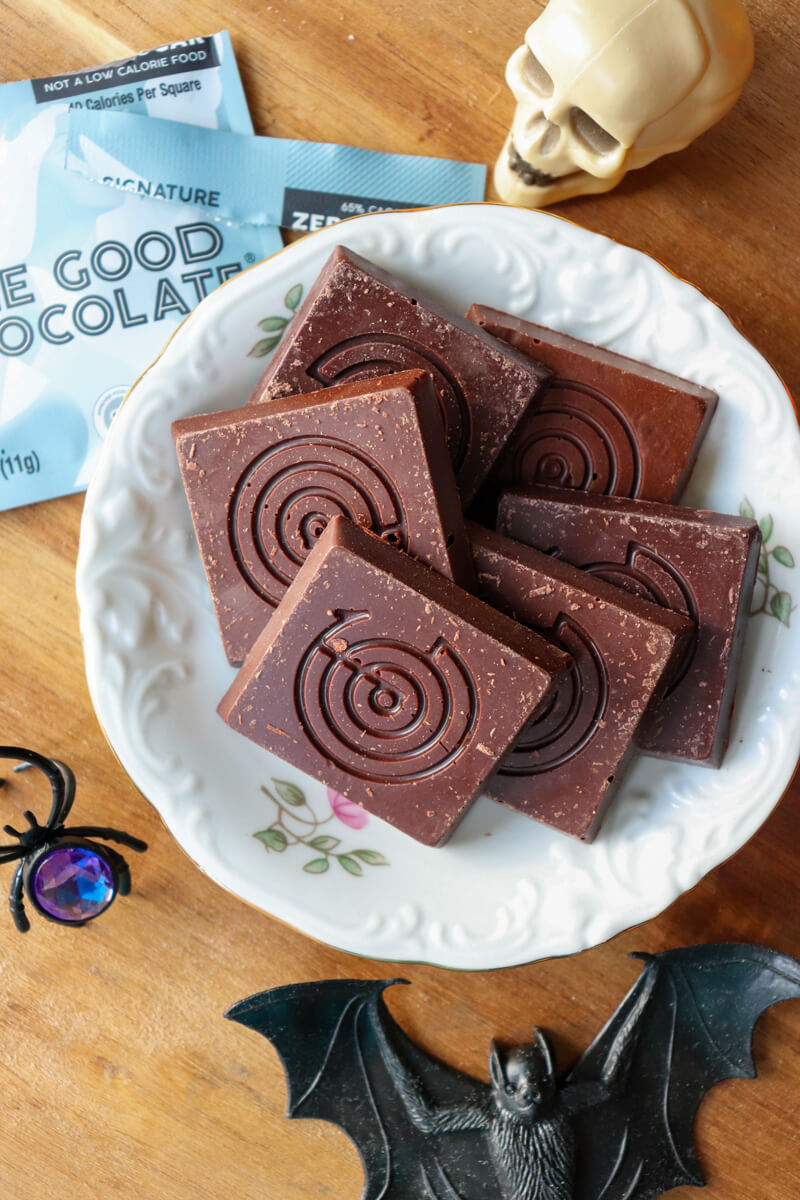 A china plate with squares of The Good Chocolate on a wooden circle with a black spider ring next to it.