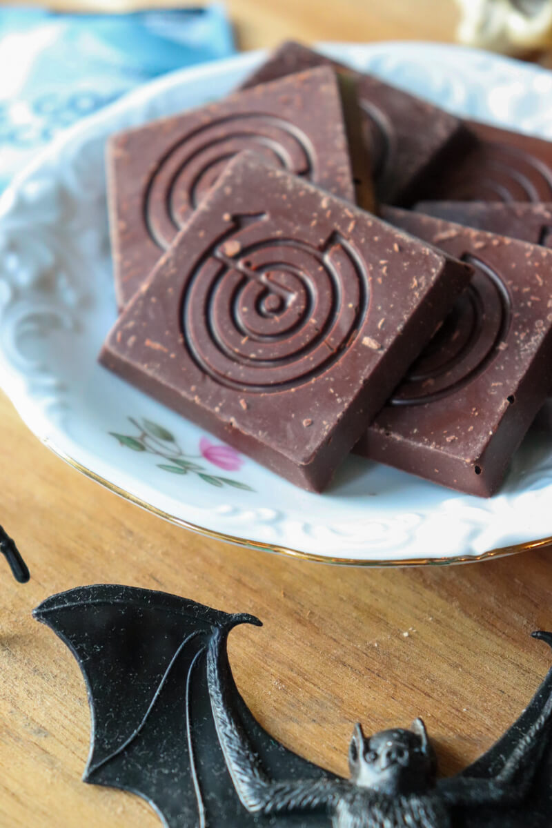 A china plate with squares of The Good Chocolate on a wooden circle with a black plastic bat next to it.