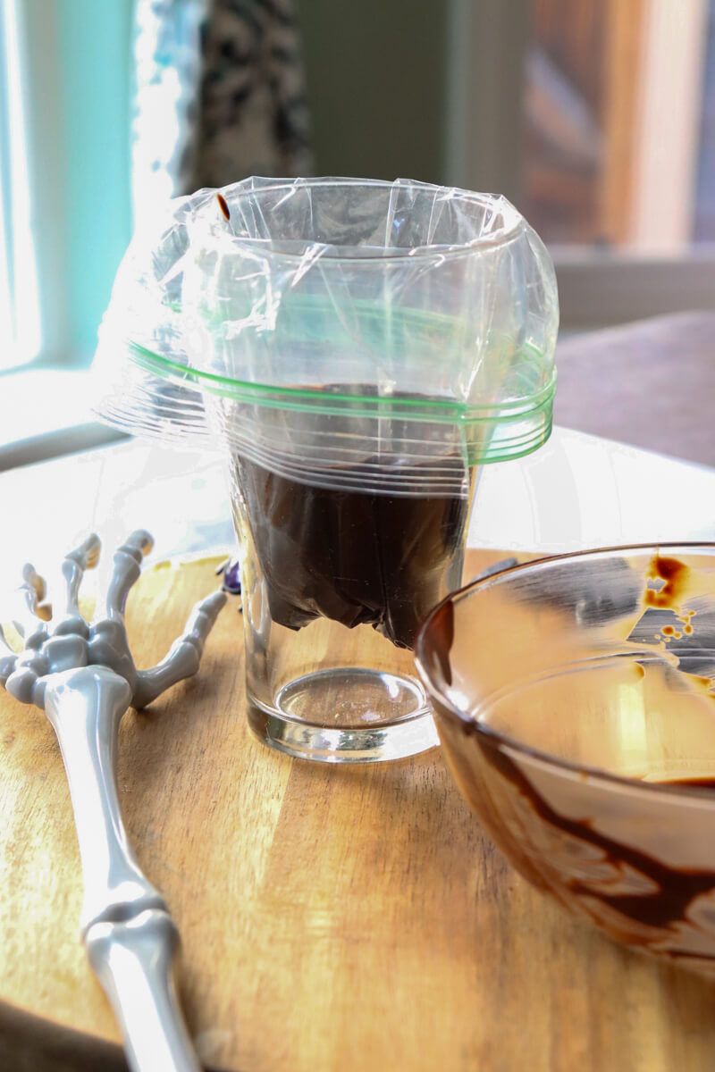 A glass with a zipper bag of melted chocolate next to a plastic skeleton hand and empty glass bowl.