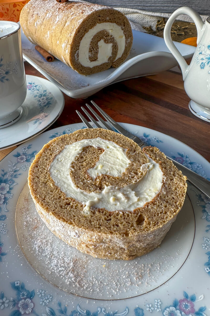 Overhead view of a piece of low carb pumpkin roll on a vintage china plate with blue flowers, partially eaten, with a fork on the plate, next to a cup of coffee and the rest of the roll