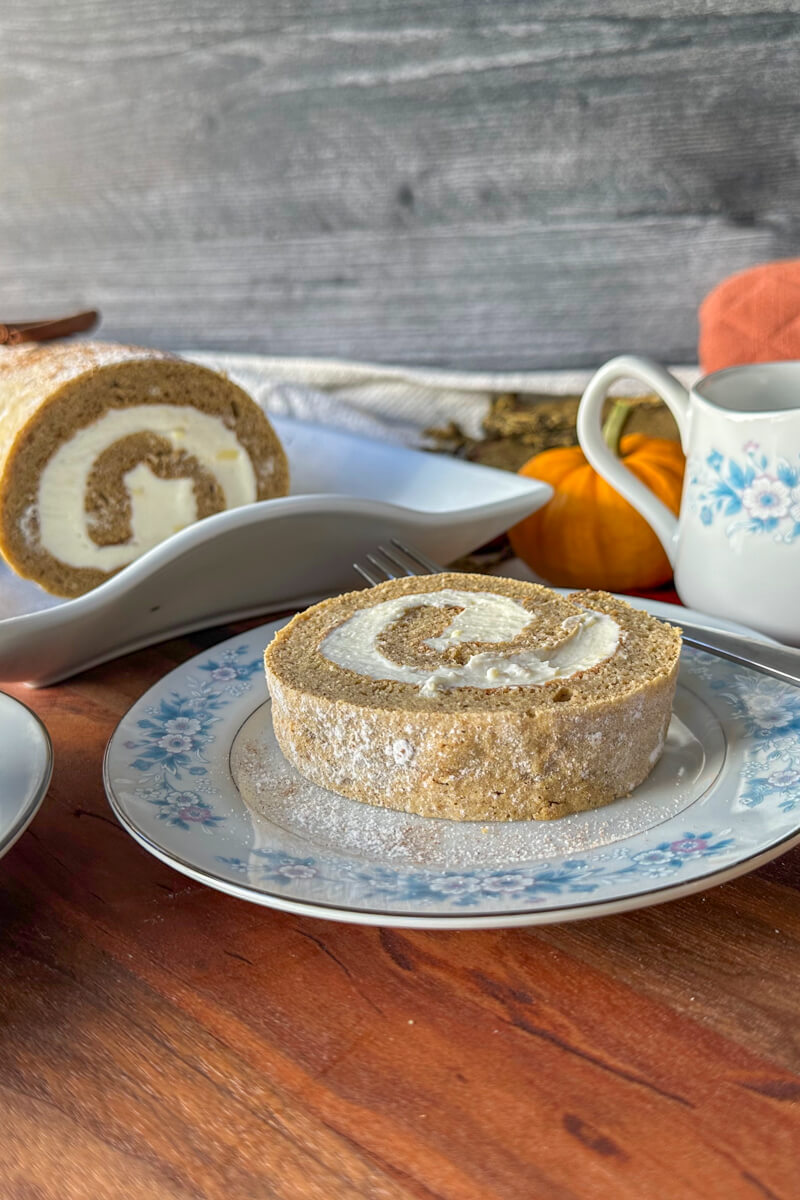 Front view of a piece of low carb pumpkin roll on a vintage china plate with blue flowers with a fork on the plate, next to a cup of coffee and the rest of the roll