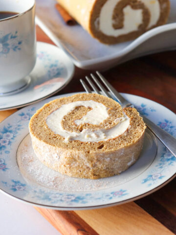 Low-carb keto pumpkin roll slice with cream cheese filling on vintage blue floral china, served with coffee and a full pumpkin roll on a wooden board in the background.